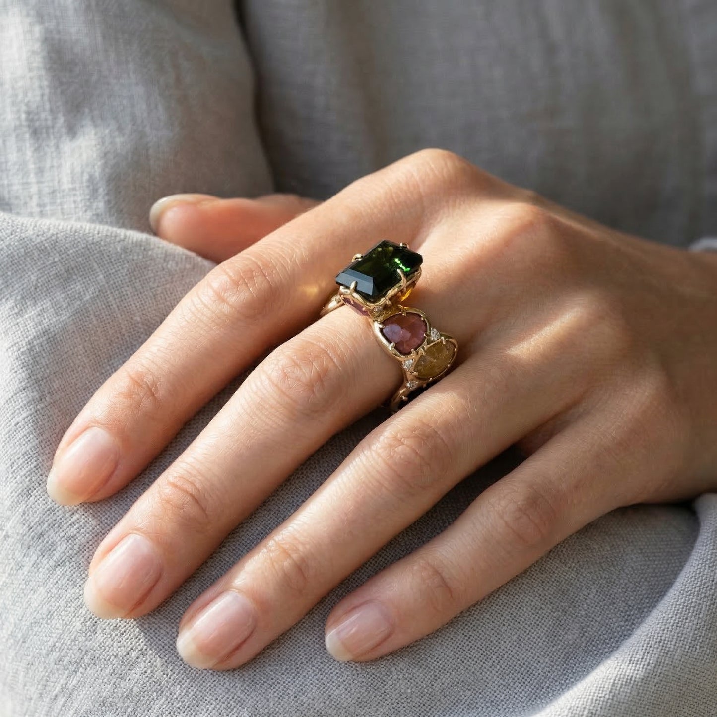 Hand wearing a gold ring with green and pink gemstones on a gray fabric background